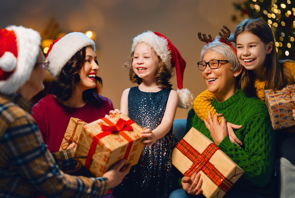 A male, female, grandma and two daughters all dressed festively holding christmas presents in front of christmas tree